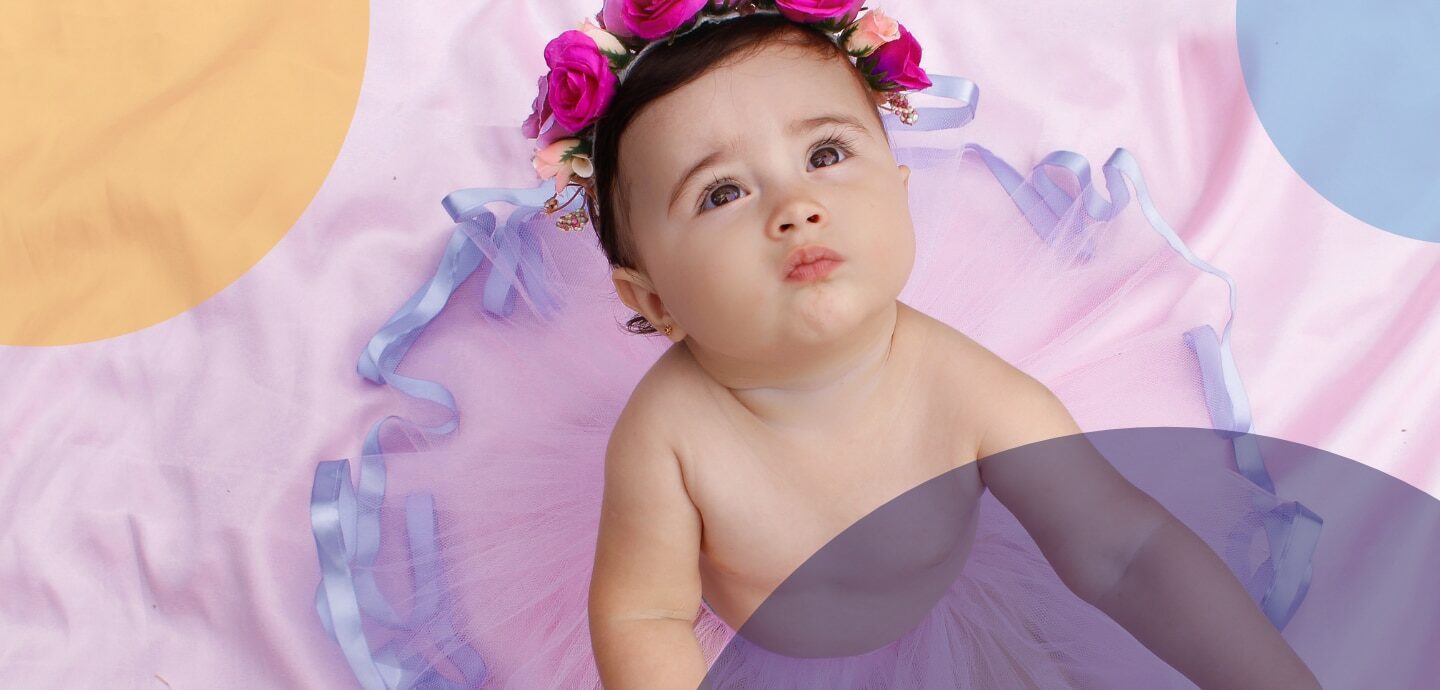 Ein Baby sitzt auf einer rosa Decke, trägt ein lila Tütü und rosafarbene Blumen im Haar.
