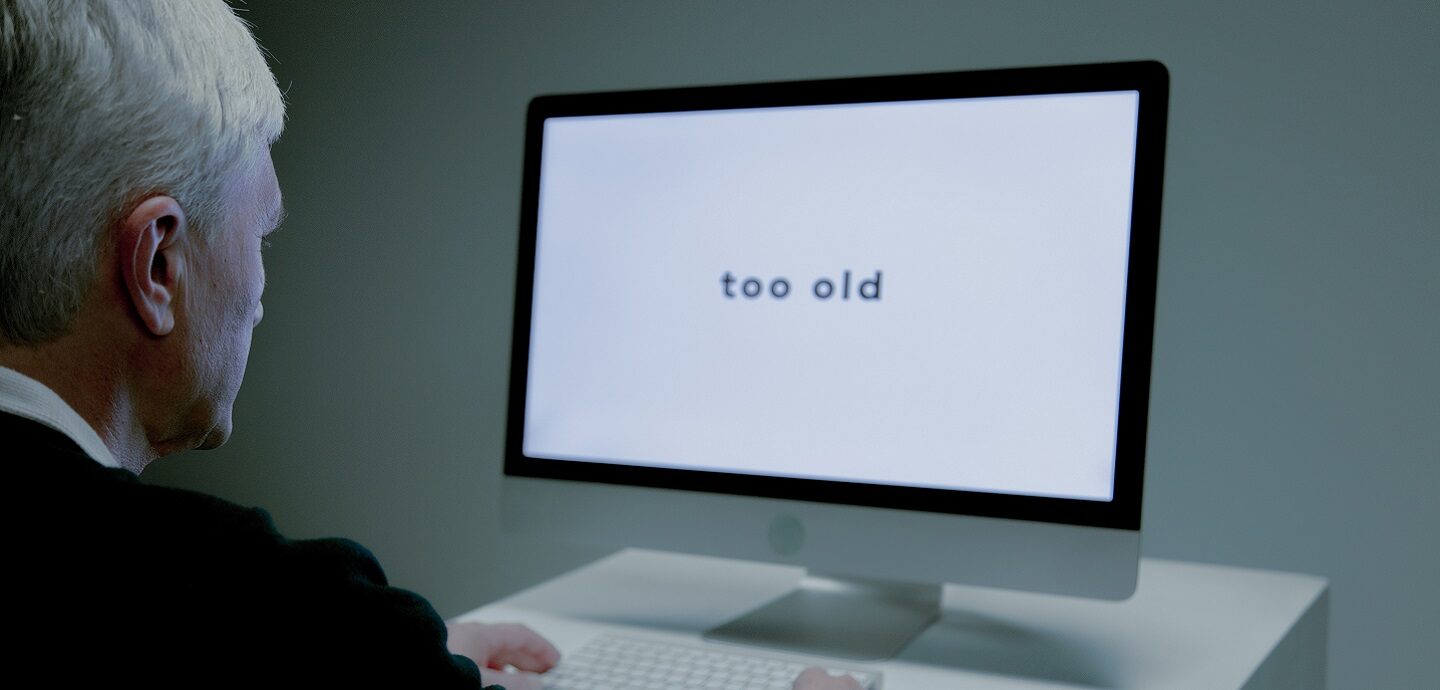 Ein Mann höheren Alters betrachtet einen Bildschirm, auf dem die Worte "too old" stehen.