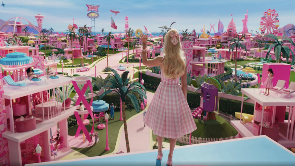 Barbie ist von hinten im Barbieland zu sehen. Sie ist eine Frau mit blonden langen Haaren und einem pinken Pünktchen-Kleid. Das Barbieland ist ebenfalls pink und ähnelt einem Freizeitpark.
