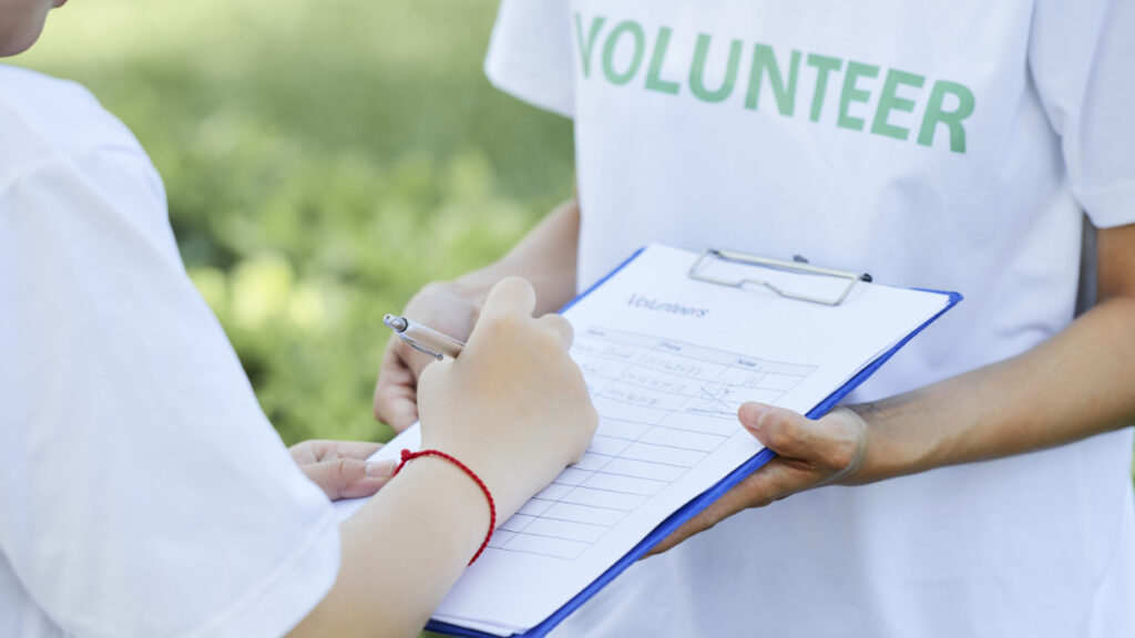 Person unterschreibt auf Klemmbrett einer Dialogerin, auf deren T-Shirt „Volunteer“ steht