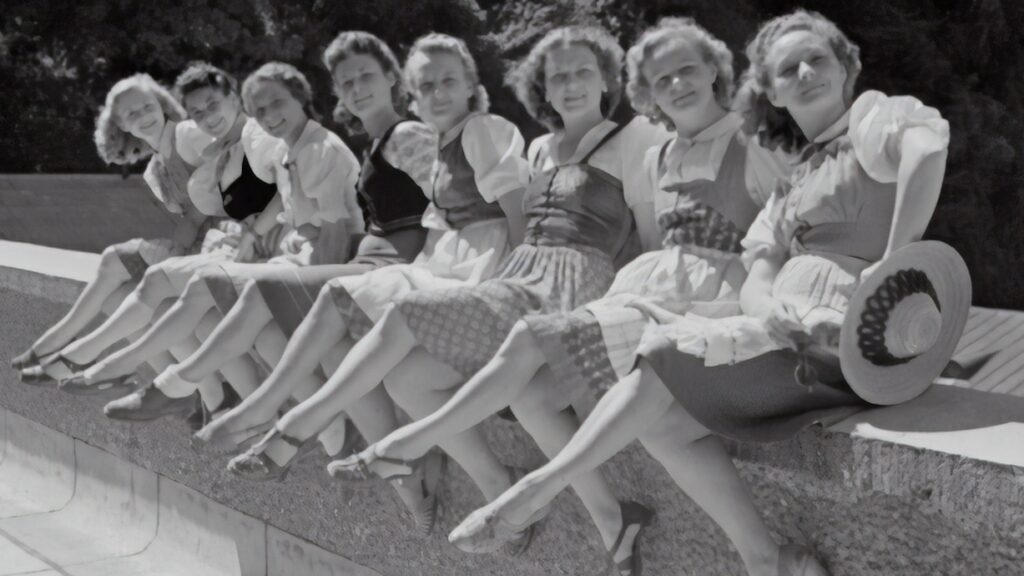 Es sind acht Frauen auf einer schwarz-weiß-Fotografie zu sehen. Sie sitzen nebeneinander auf einer Mauer und tragen alle Kleider, die auf Kniehöhe aufhören. Sie blicken in die Kamera.