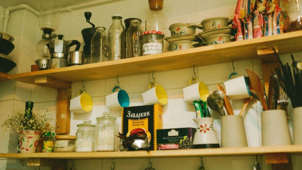 einfaches Küchenregal vor vergilbter Wand mit provisorischer Aufhängung für Tassen. Tütensuppen und Küchenutensilien sind dicht an dicht darauf geräumt.