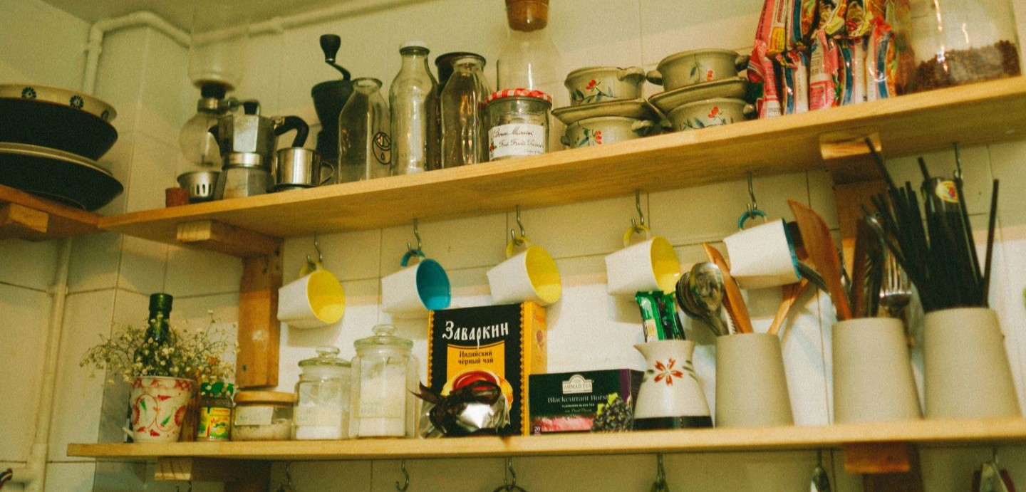 einfaches Küchenregal vor vergilbter Wand mit provisorischer Aufhängung für Tassen. Tütensuppen und Küchenutensilien sind dicht an dicht darauf geräumt.
